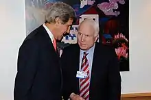 Two white men in suits standing next to each other inside a room with white walls lit by a camera flash. The one on the left has his right side to the viewer and is taller; the one on the right is older, with thin white hair, and faces the viewer
